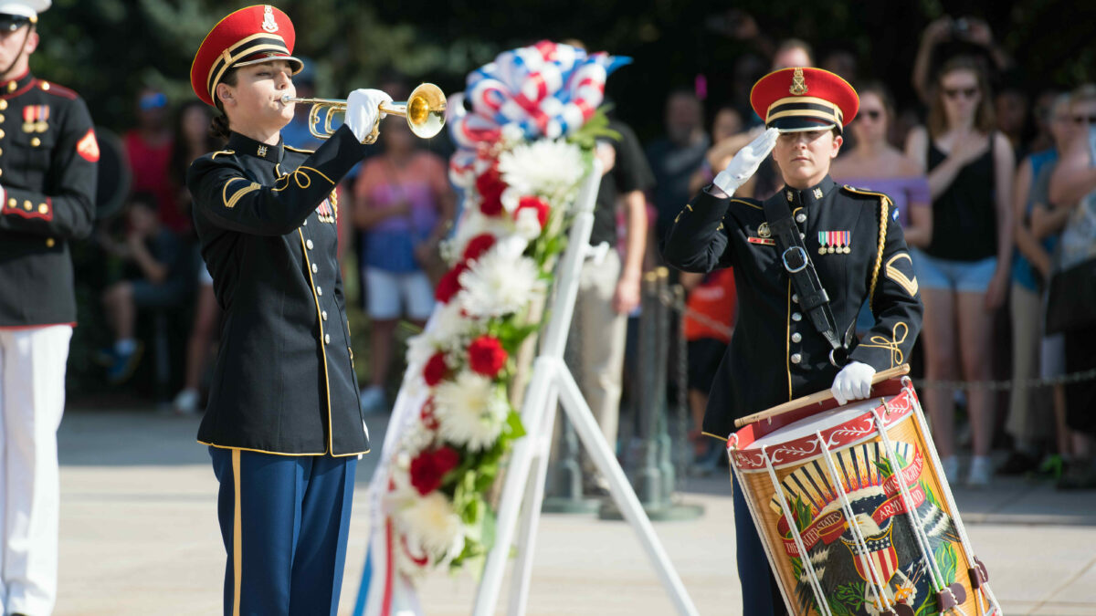 A bugler performs Taps at the Tomb of the Unknown Soldier. A wreath is present and a drummer is saluting.